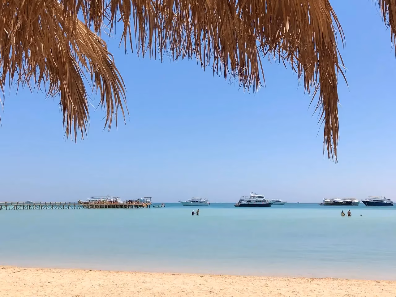 Een palmdak boven een zandstrand met helder water en een pier in de verte