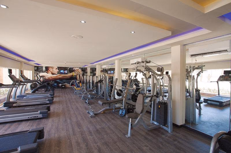 Fitnessruimte met loopbanden, krachtapparaten en een spiegelwand in een oudere inrichting met een paarse lichtlijn in Sindbad Club in Hurghada