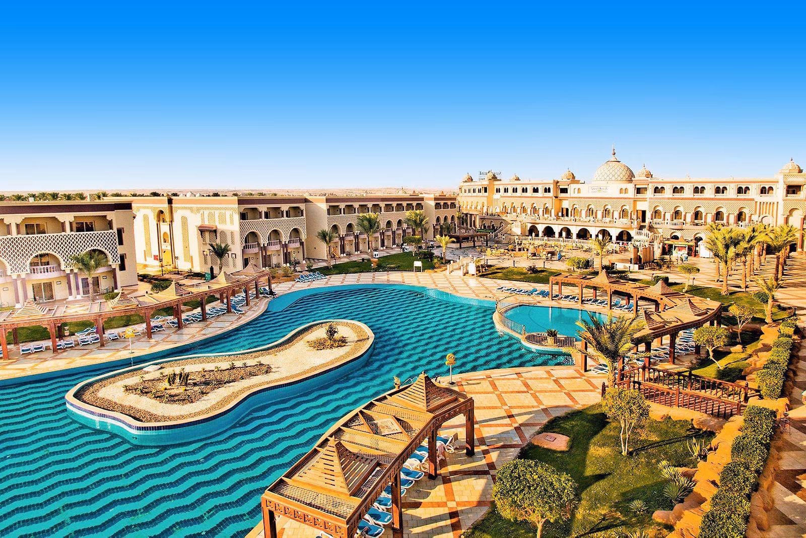 Zwembad met golvend blauw tegelpatroon en een zandeiland, houten pergola's met ligbedden en een paleisachtig hoofdgebouw met witte koepels bij Sentido Mamlouk Palace Resort in Hurghada
