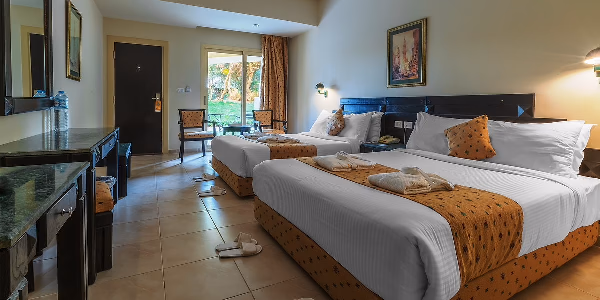 Familiekamer met twee tweepersoonsbedden met oranje bedlopers en een tuindeur naar een groen terras, in oudere inrichting, in Sindbad Club in Hurghada