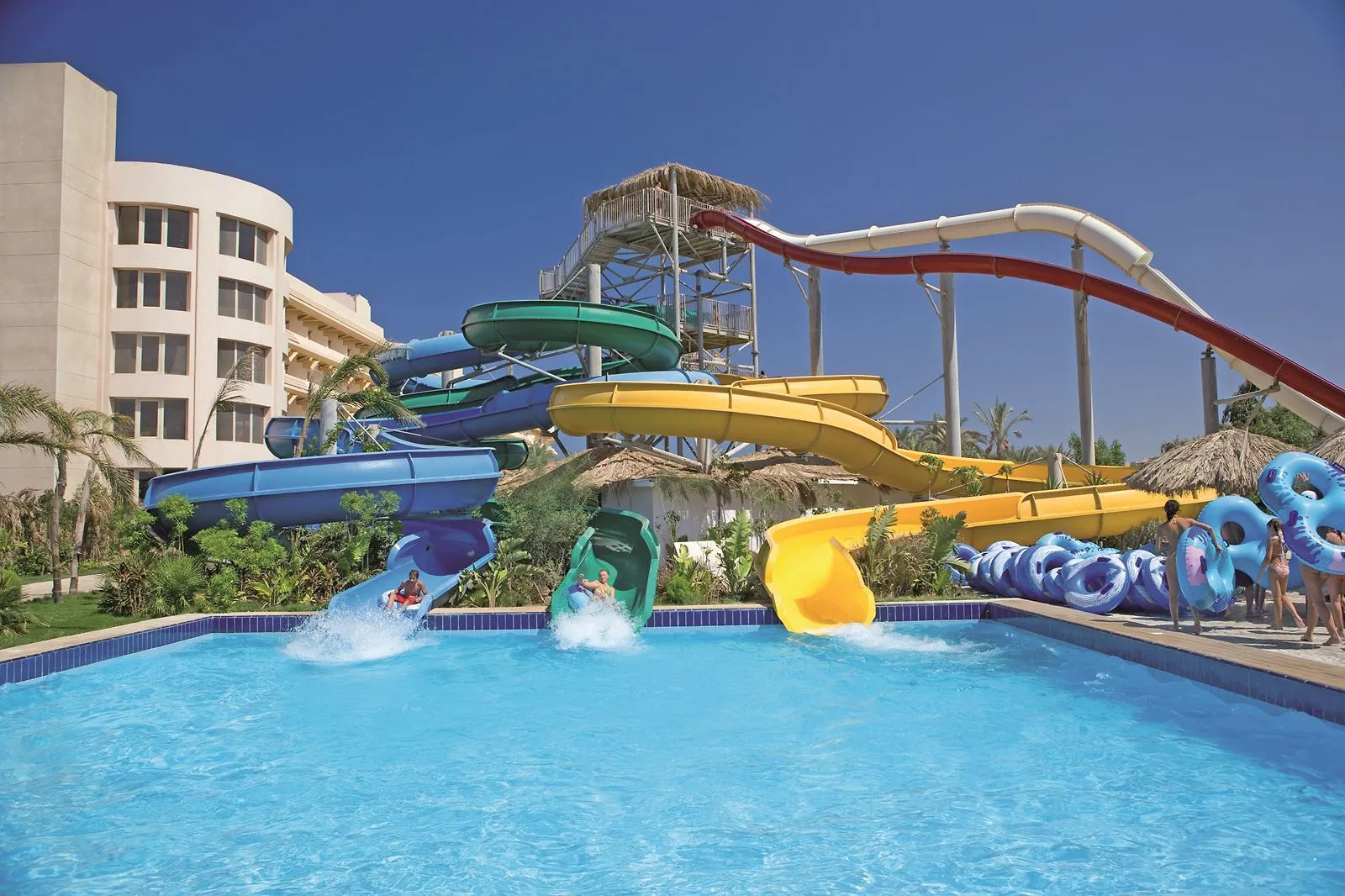 Blauwe, groene, gele en rode glijbanen vanaf een toren met rieten dak die uitkomen in het landingsbad van het aquapark van Sindbad Club in Hurghada