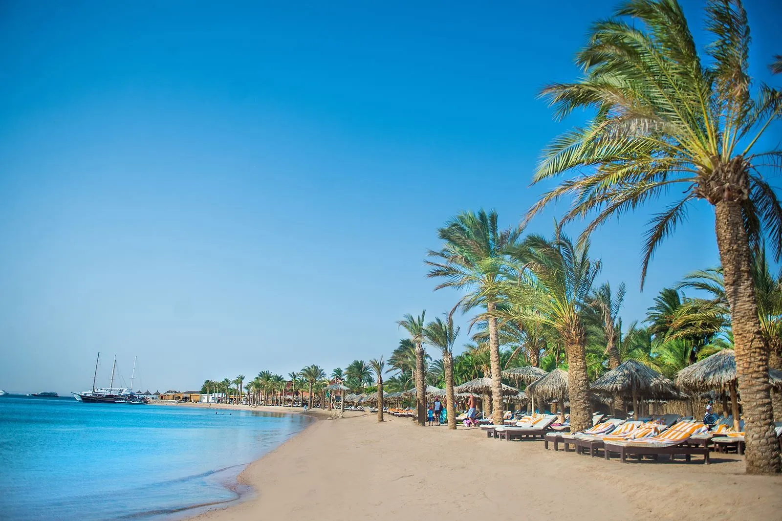 Lang zandstrand met palmen, rieten parasols en ligbedden met gestreepte kussens aan een rustige zee met zeilschepen bij Sindbad Club in Hurghada