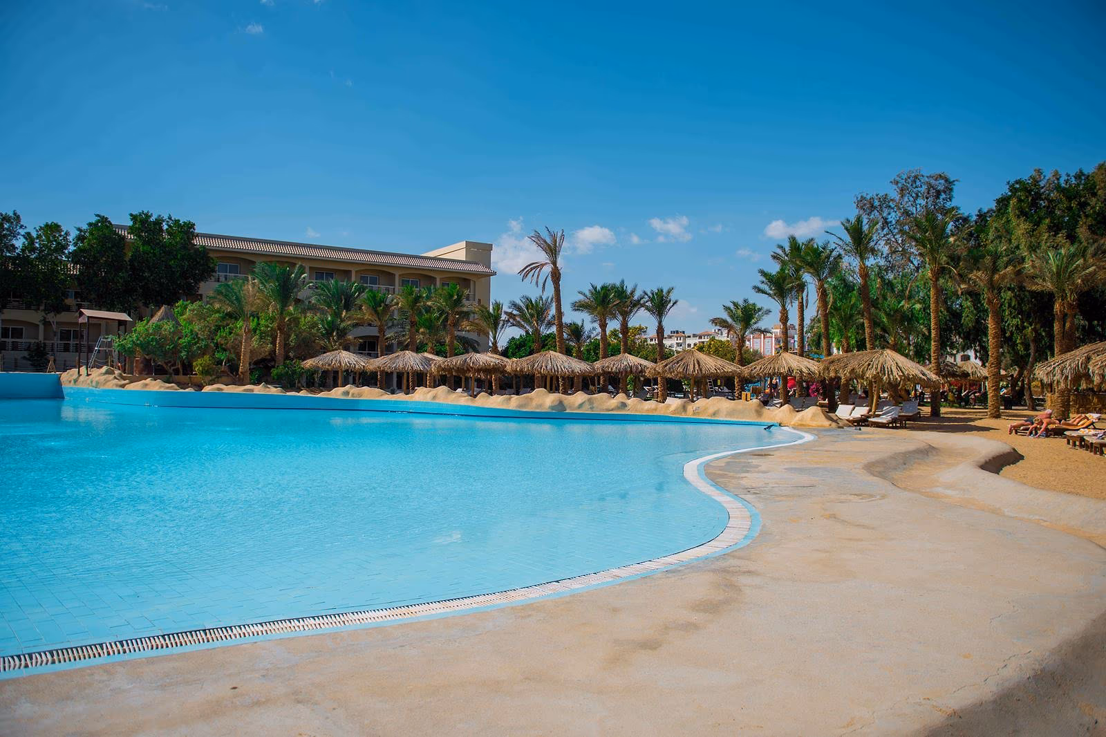 Zwembad met een flauw aflopende strandinstap tussen palmen en rieten parasols, met een hotelvleugel op de achtergrond, bij Sindbad Club in Hurghada