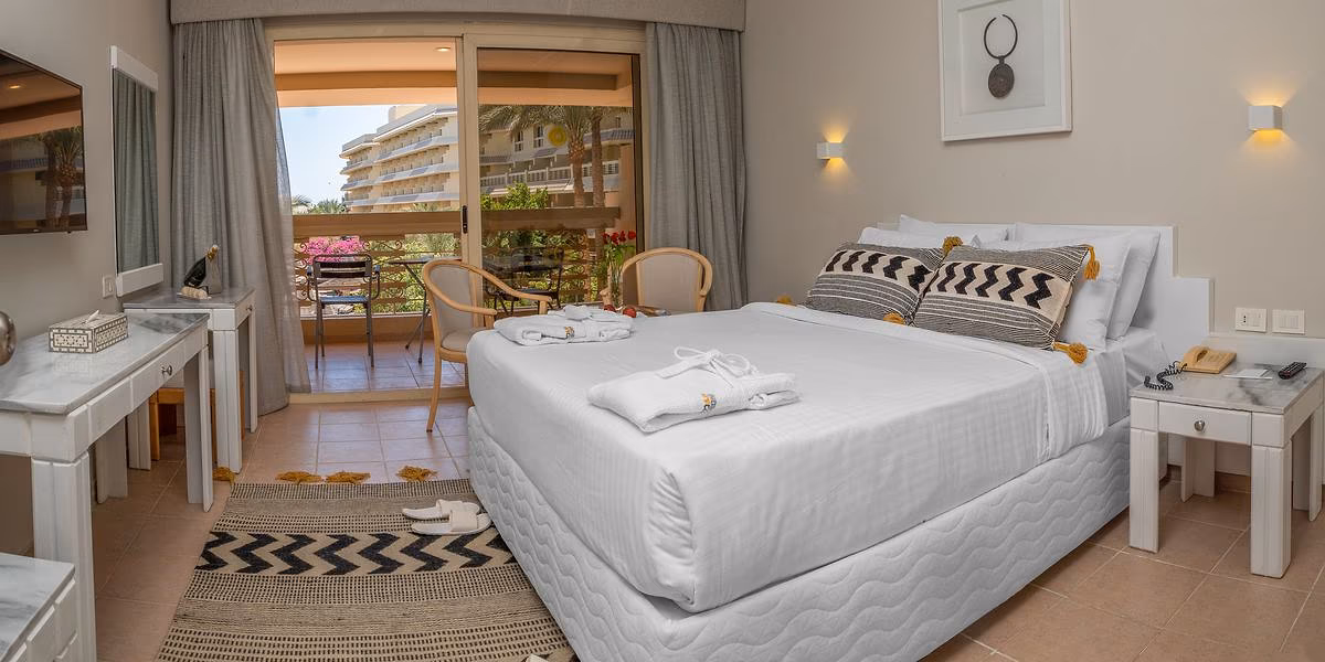 Kamer met een tweepersoonsbed met zwart-wit sierkussens en een balkon met zitje en uitzicht op een hotelvleugel met palmen in Sindbad Club in Hurghada