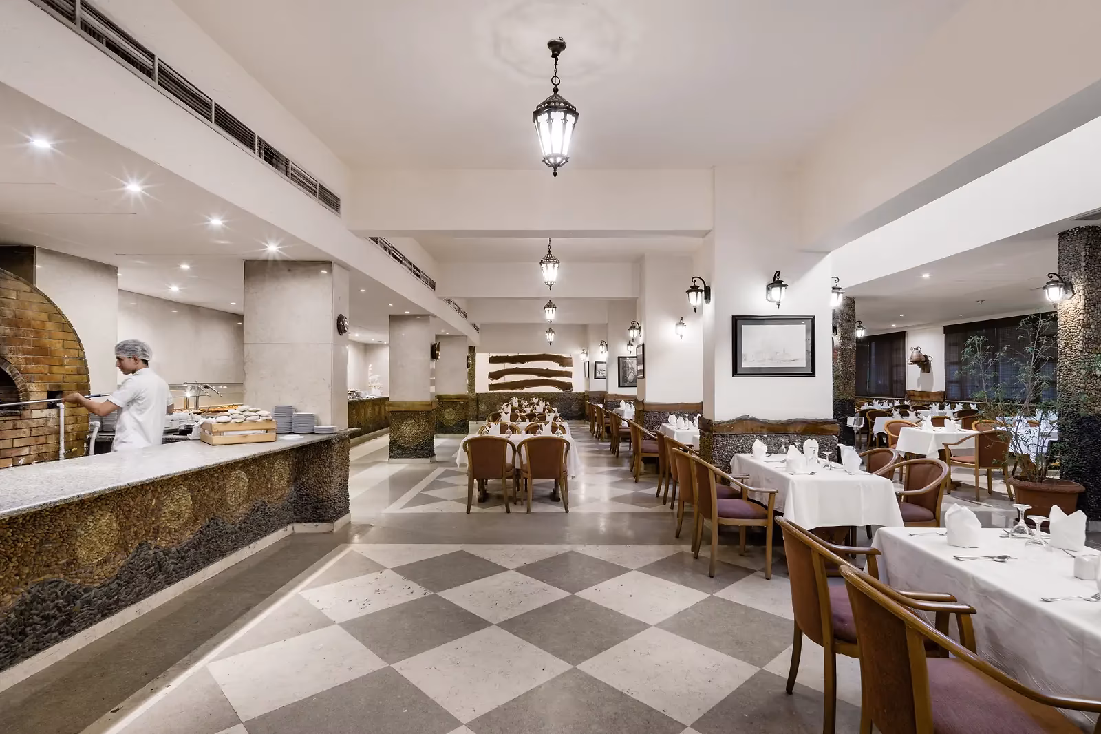 Restaurantzaal met een zwart-wit geblokte vloer, wit gedekte tafels en een kok bij een stenen oven in Sindbad Club in Hurghada