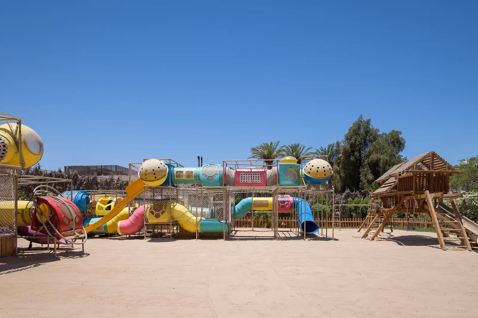 Grote speeltuin op zand met een kleurig buizenparcours, gele en blauwe glijbaantjes en een houten speelhuis tussen palmen bij Sindbad Club in Hurghada