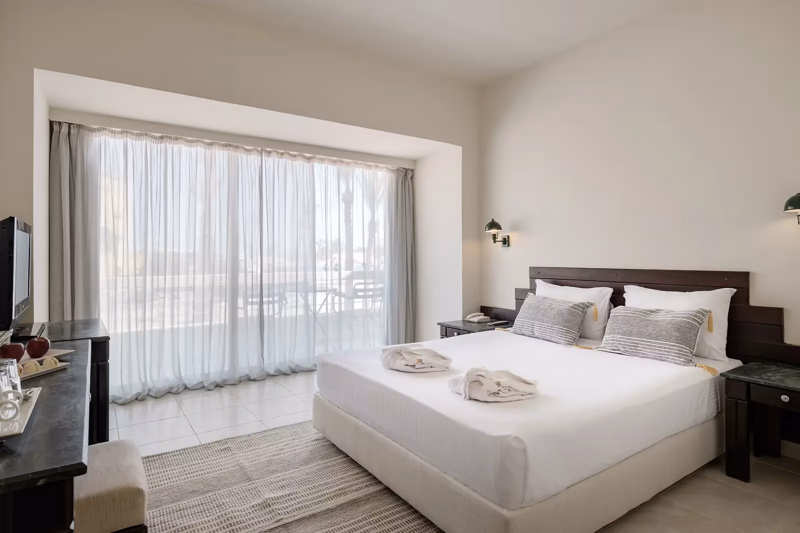 Kamer met een kingsize bed met gestreepte kussens, donkere nachtkastjes en witte vitrage voor een glazen pui naar buiten in Sindbad Club in Hurghada