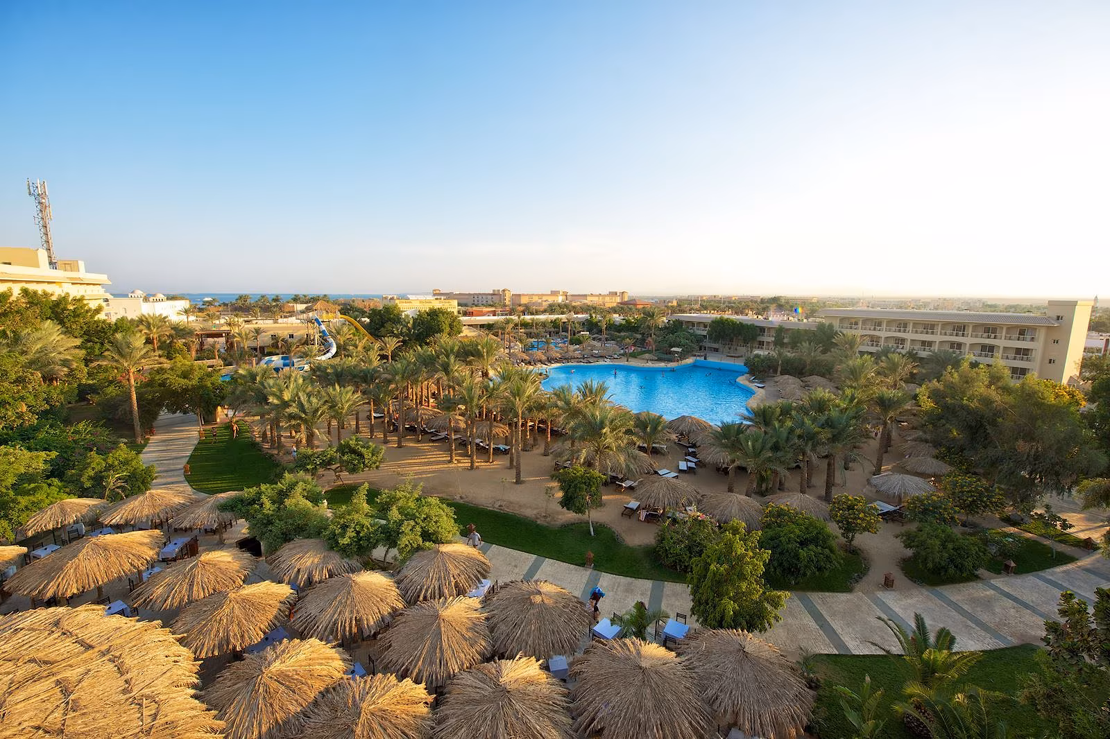 Luchtfoto van de tuin met rieten parasols, paden, gazons en het grote zwembad van Sindbad Club in Hurghada, met de glijbaantoren links en de zee aan de horizon