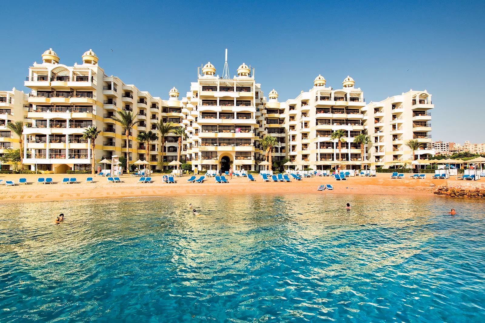 Witte hotelgebouwen met koepels van Sunrise Holidays Resort in Hurghada aan een kleine baai met zandstrand, blauwe ligbedden en helder water